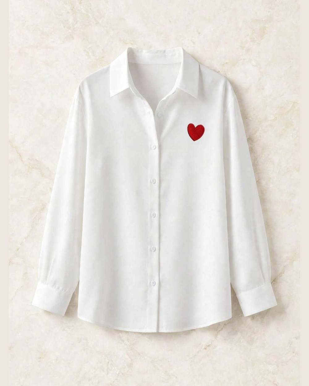 Camisa blanca de botones con corazón rojo bordado, aislada (gancho, carterita y círculo 'EMBROIDERY' eliminados) sobre mármol marfil