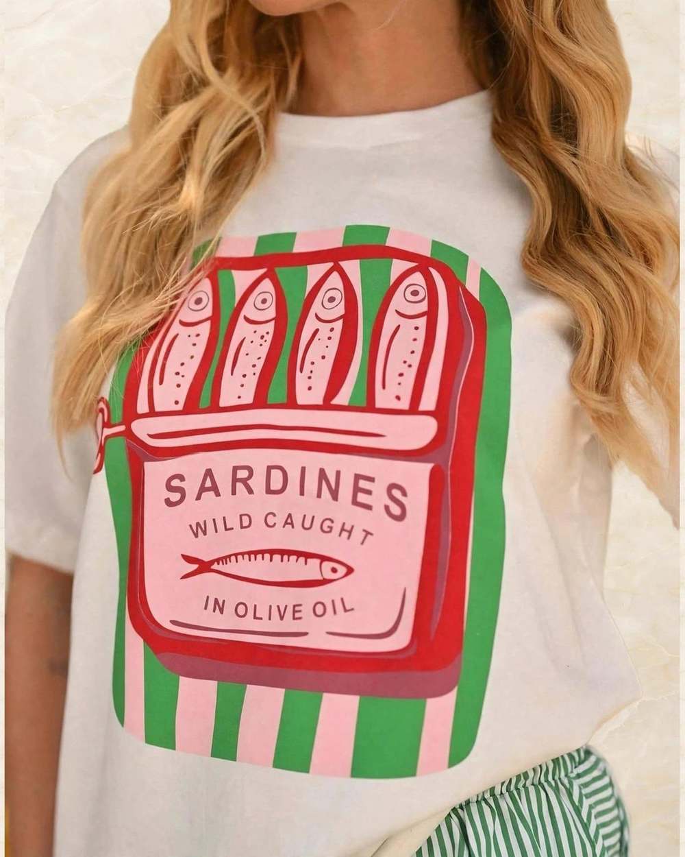 Franela blanca con estampado 'SARDINES' en verde y rosa., aislado sobre mármol marfil