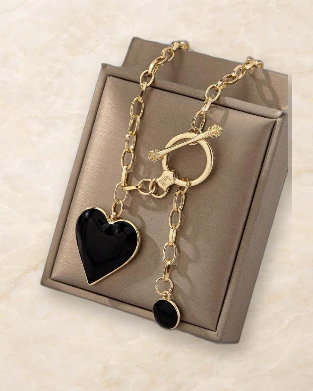Collar de cadena dorada con dije de corazon negro y colgante redondo negro., aislado sobre mármol marfil