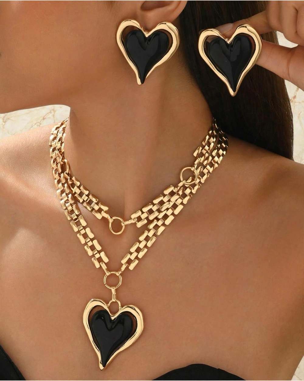 Set de collar de cadena dorada en capas con dije de corazon negro y zarcillos de corazon negro a juego., aislado sobre mármol marfil