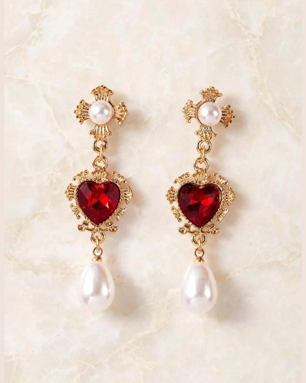 Zarcillos: dije de cruz dorada con perla, corazón rojo facetado y gota de perla, aislados sobre mármol marfil
